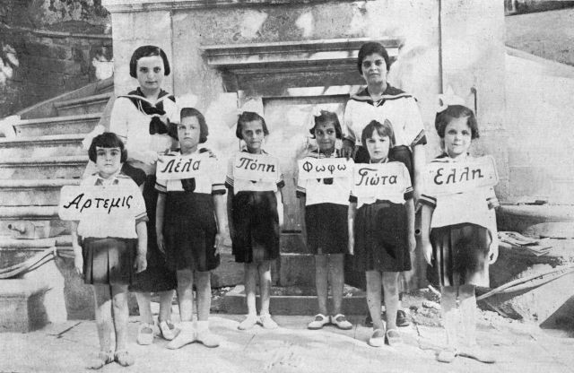 Οι σπουδάστριες τού Αρσακείου Διδασκαλείου Πατρών, στο πλαίσιο τής πρακτικής εξάσκησής τους, διδάσκουν ορθογραφία στις μικρές μαθήτριες τού Δημοτικού. Αυτό δικαιολογεί και το σχεδόν ομοιόμορφο τής στολής των σπουδαστριών με τις μαθήτριες. Η ναυτική στολή των μαθητριών με τους χαρακτηριστικούς φιόγκους στο κεφάλι είναι εντυπωσιακή, όπως και το στήσιμο τής φωτογραφίας. E032a